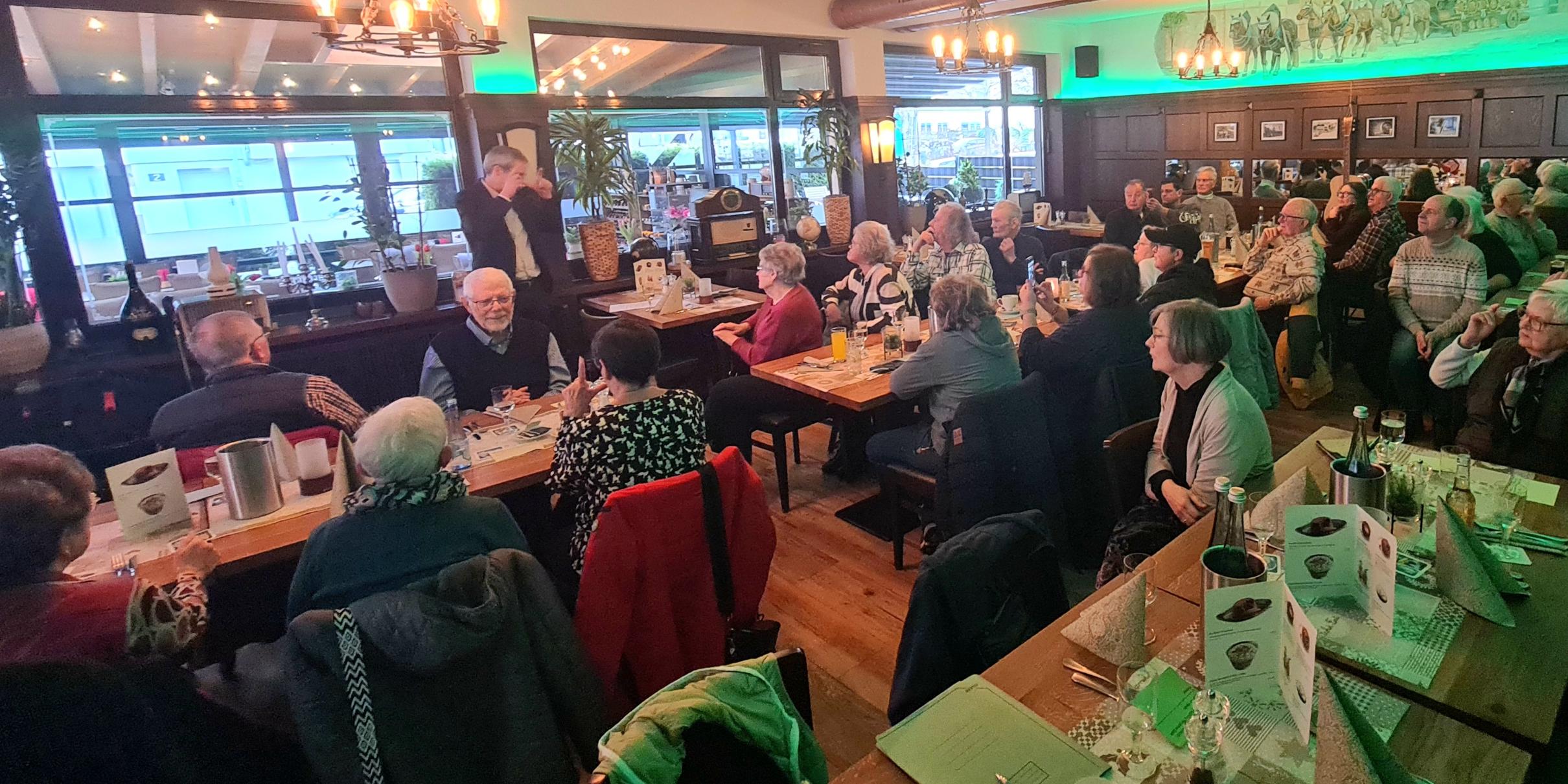 Treffen in einem Restaurant: Gruppe sitzt an Tischen und hört einem stehenden Redner im Gastraum zu.