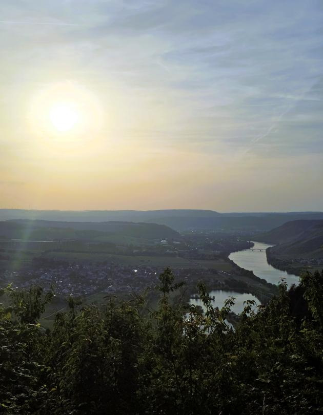 Blick von einem Hang auf ein Flusstal mit Ortschaften; die tief stehende Sonne beleuchtet die Landschaft.