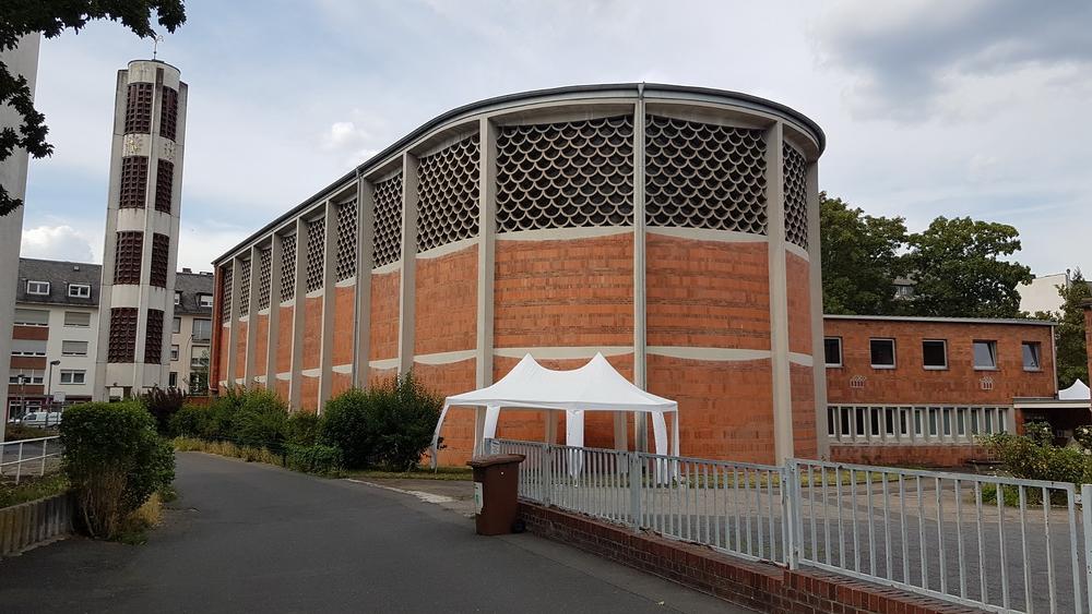 Moderne Backsteinkirche mit freistehendem Turm, Vorplatz und weißem Pavillon unter bewölktem Himmel. (c) Ralf Schmitz Moderne Backsteinkirche mit freistehendem Turm, Vorplatz und weißem Pavillon unter bewölktem Himmel.