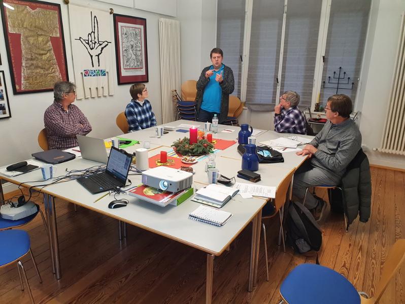 Sitzung in einem Besprechungsraum: Fünf Personen am Tisch, eine Person präsentiert stehend mit Gebärden. (c) Ralf Schmitz Sitzung in einem Besprechungsraum: Fünf Personen am Tisch, eine Person präsentiert stehend mit Gebärden.