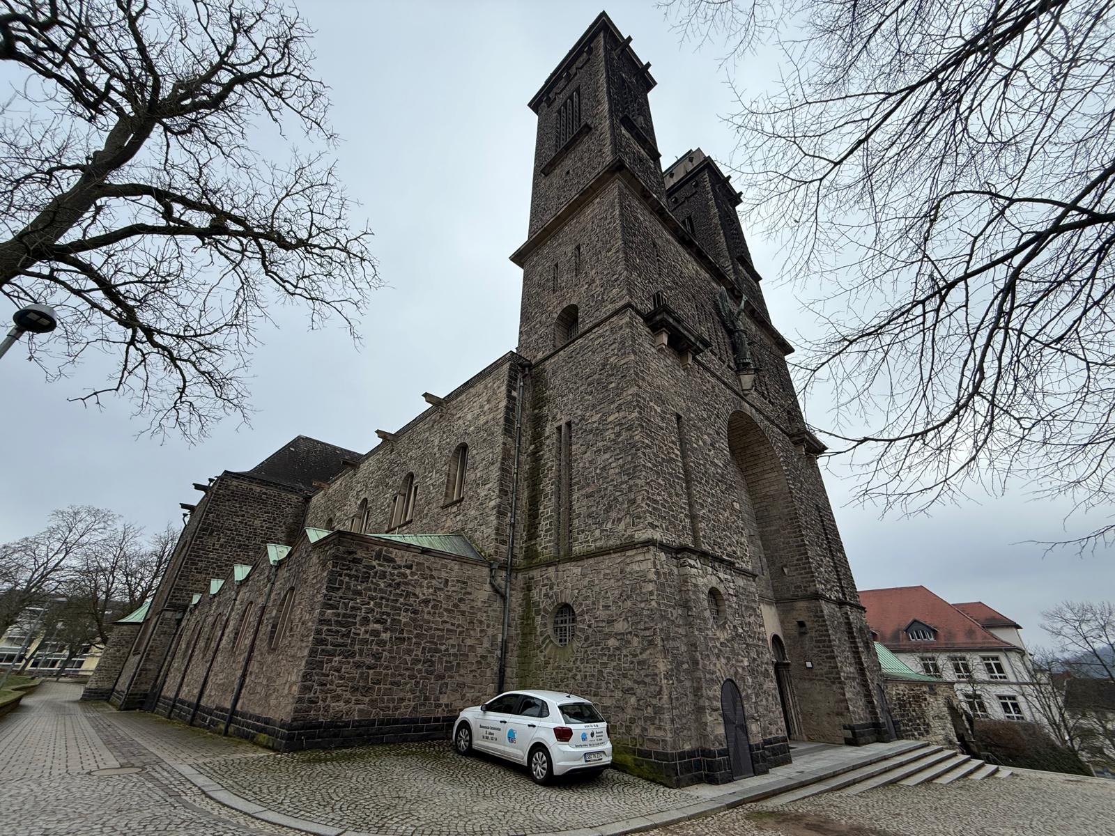 Große Kirche aus Naturstein mit markantem Turm, aufgenommen von einem gepflasterten Vorplatz.