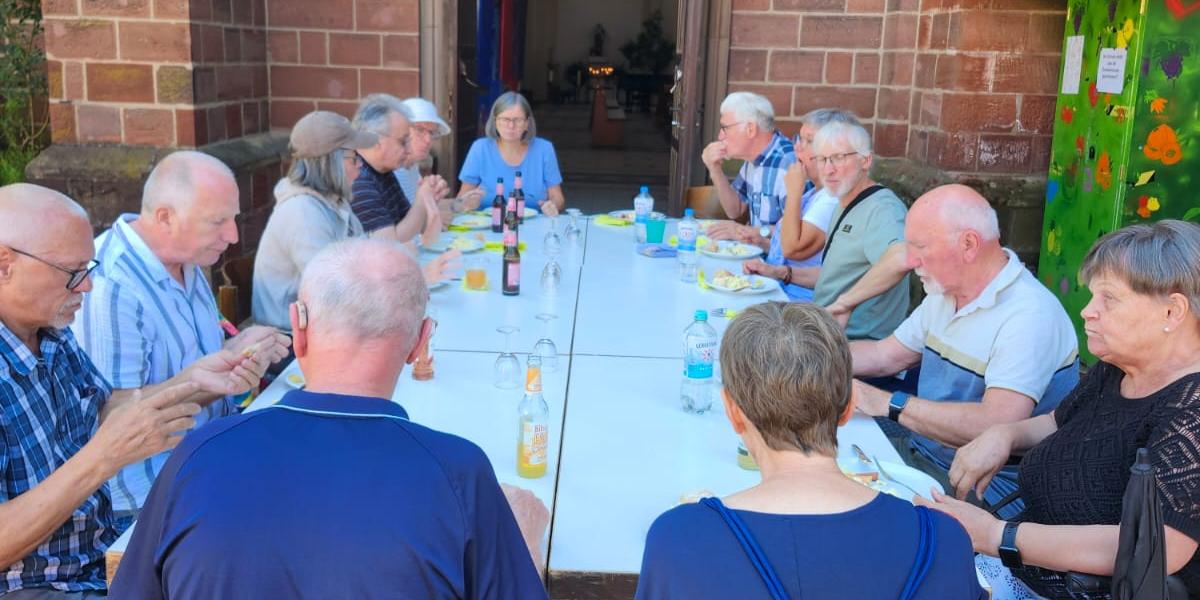 Menschen sitzen an einer langen Tafel vor der Kirche und essen gemeinsam im Freien.