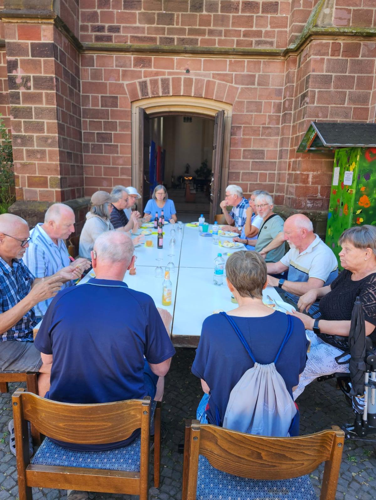 Menschen sitzen an einer langen Tafel vor der Kirche und essen gemeinsam im Freien.