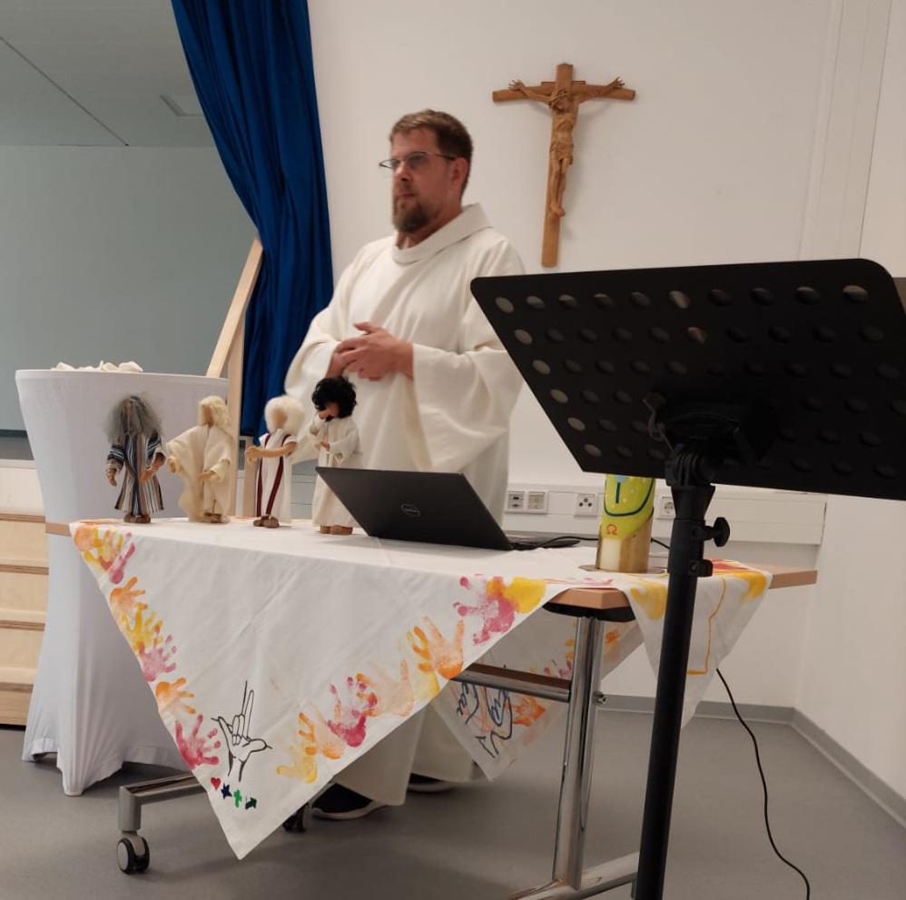 Person in liturgischer Kleidung präsentiert vor einem Tisch mit biblischen Darstellungen und Kruzifix.