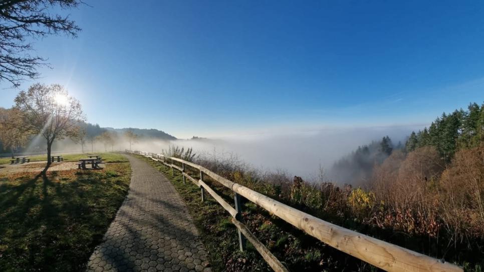Weg mit Holzgeländer führt zu einem Aussichtspunkt über einem nebelverhangenen Tal unter blauem Himmel.