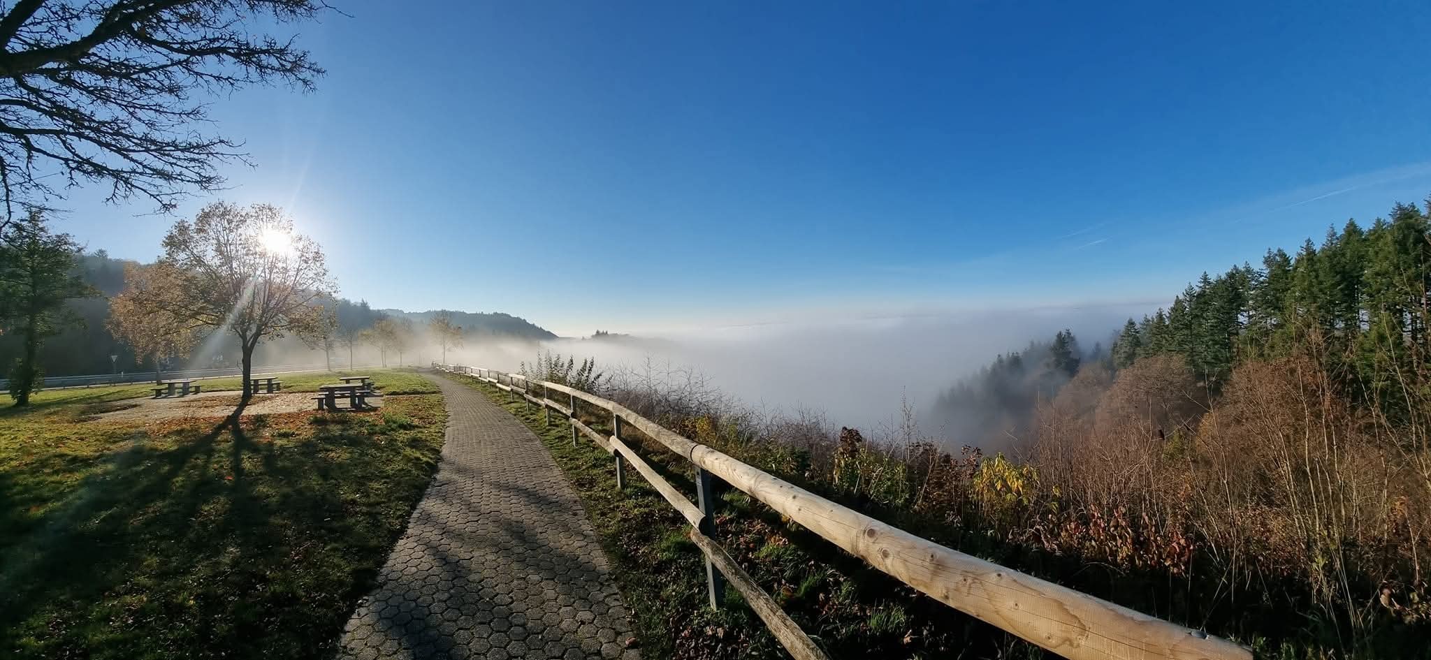 Weg mit Holzgeländer führt zu einem Aussichtspunkt über einem nebelverhangenen Tal unter blauem Himmel.