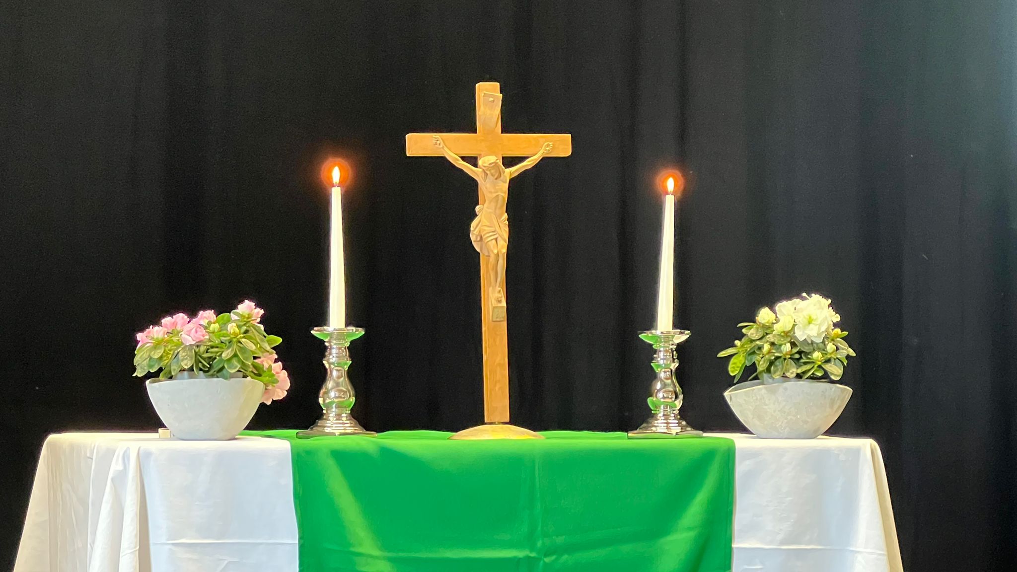 Ein Altar mit einem Holzkreuz in der Mitte, dazu 2 Kerzen und 2 Blumentöpfe.