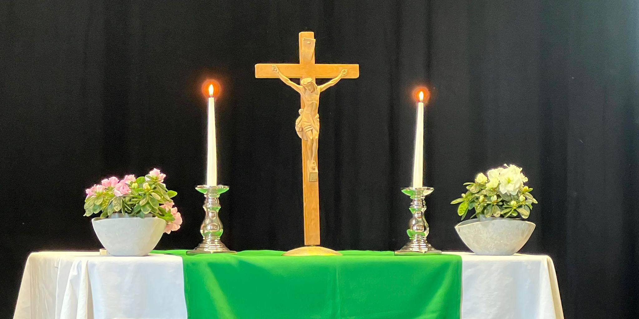 Ein Altar mit einem Holzkreuz in der Mitte, dazu 2 Kerzen und 2 Blumentöpfe.