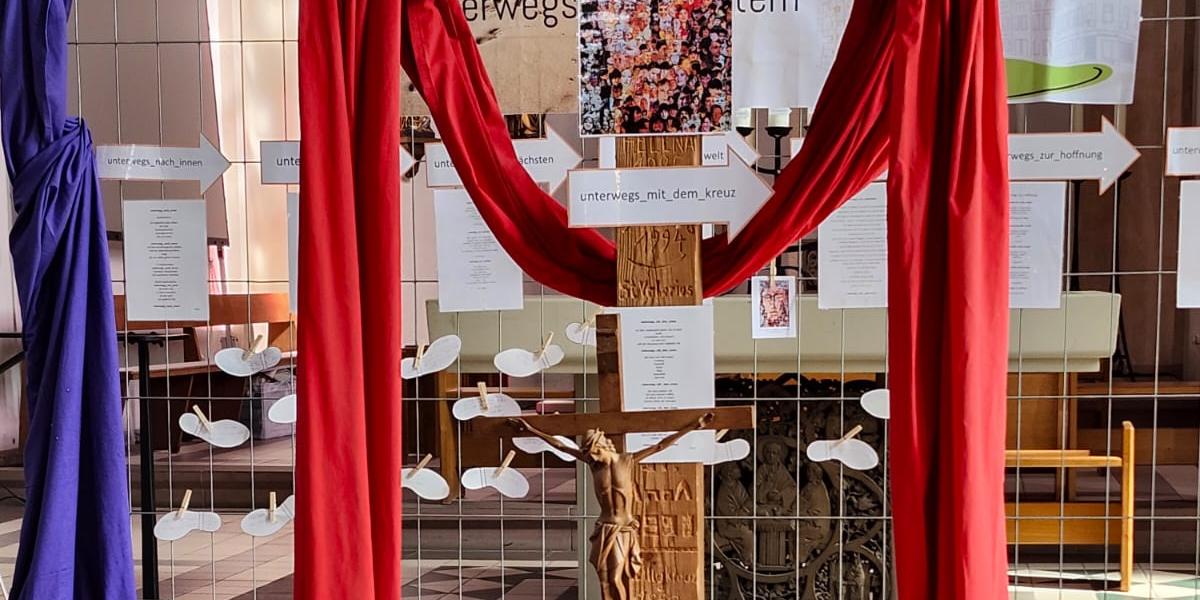 Holzkreuz mit roten Tüchern, Kerzen und Blumen im Altarraum einer Kirche.