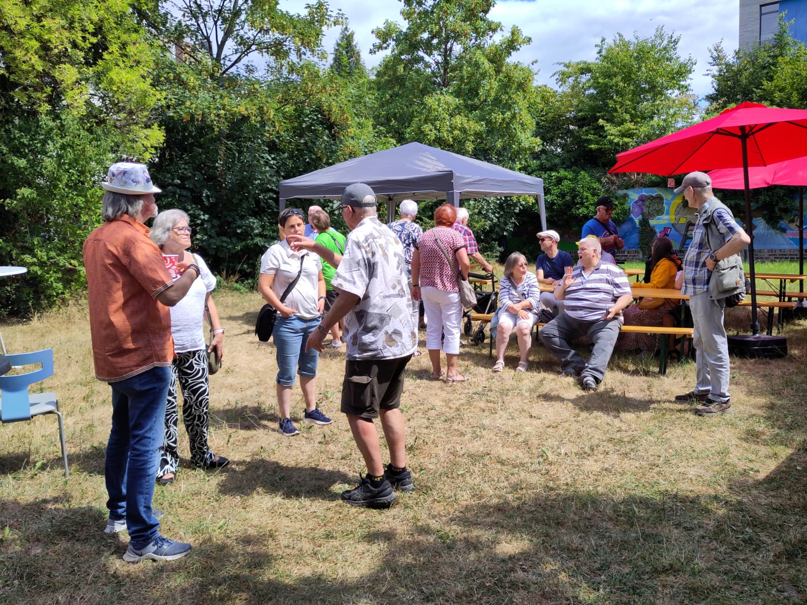 Menschen unter einem Pavillon und Sonnenschirmen bei einem sommerlichen Treffen im Freien im Gespräch.