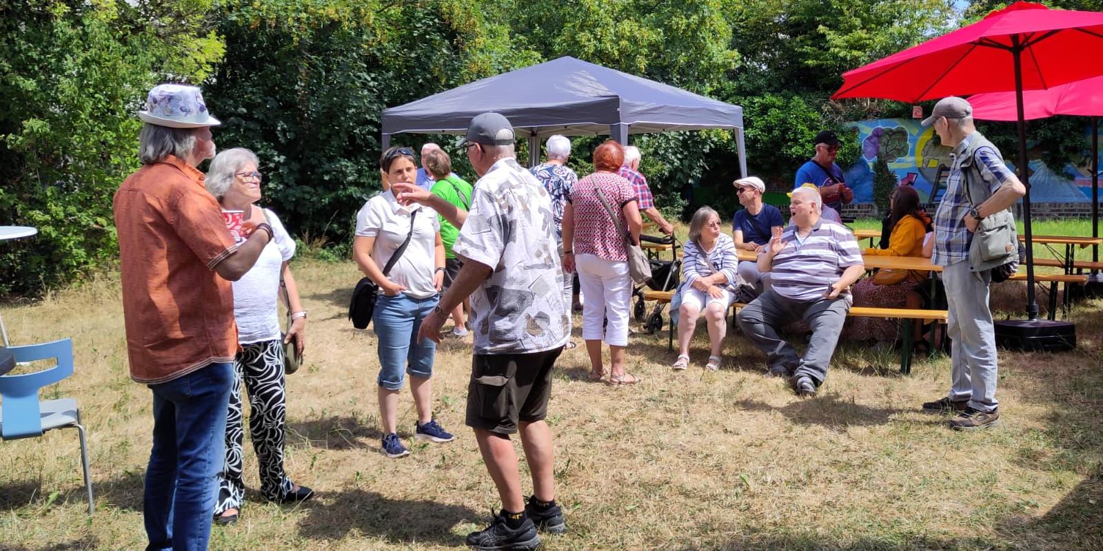 Menschen unter einem Pavillon und Sonnenschirmen bei einem sommerlichen Treffen im Freien im Gespräch.