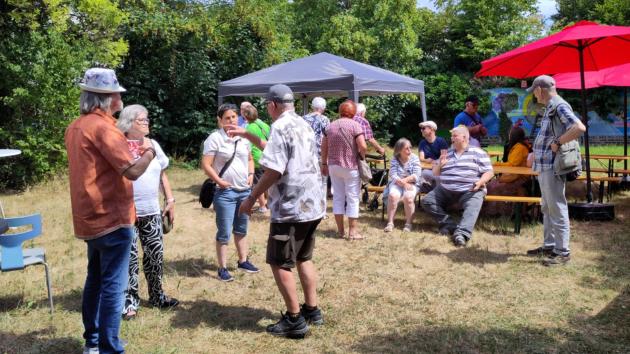 Menschen unter einem Pavillon und Sonnenschirmen bei einem sommerlichen Treffen im Freien im Gespräch.