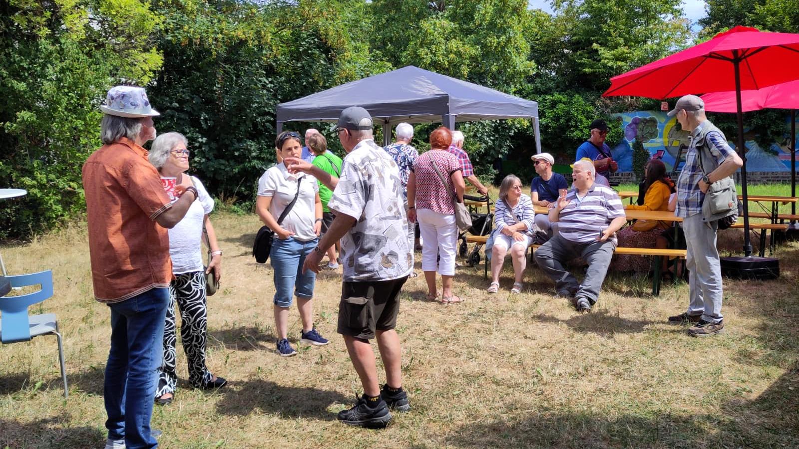 Menschen unter einem Pavillon und Sonnenschirmen bei einem sommerlichen Treffen im Freien im Gespräch.