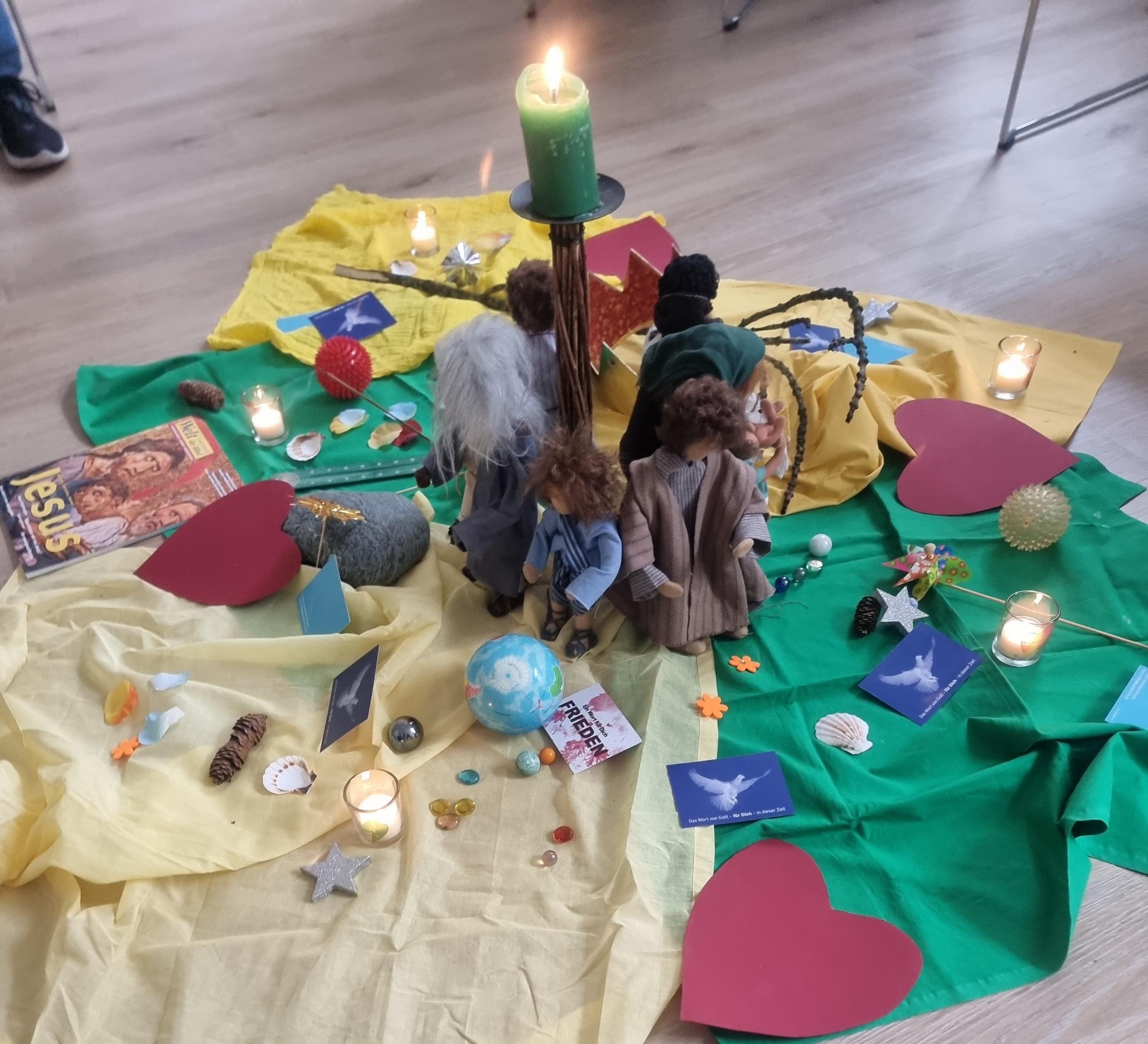 Bodenbild mit Kerze, Figuren, Herzen und Glaubenssymbolen zur gemeinsamen Andacht oder Gruppenarbeit.