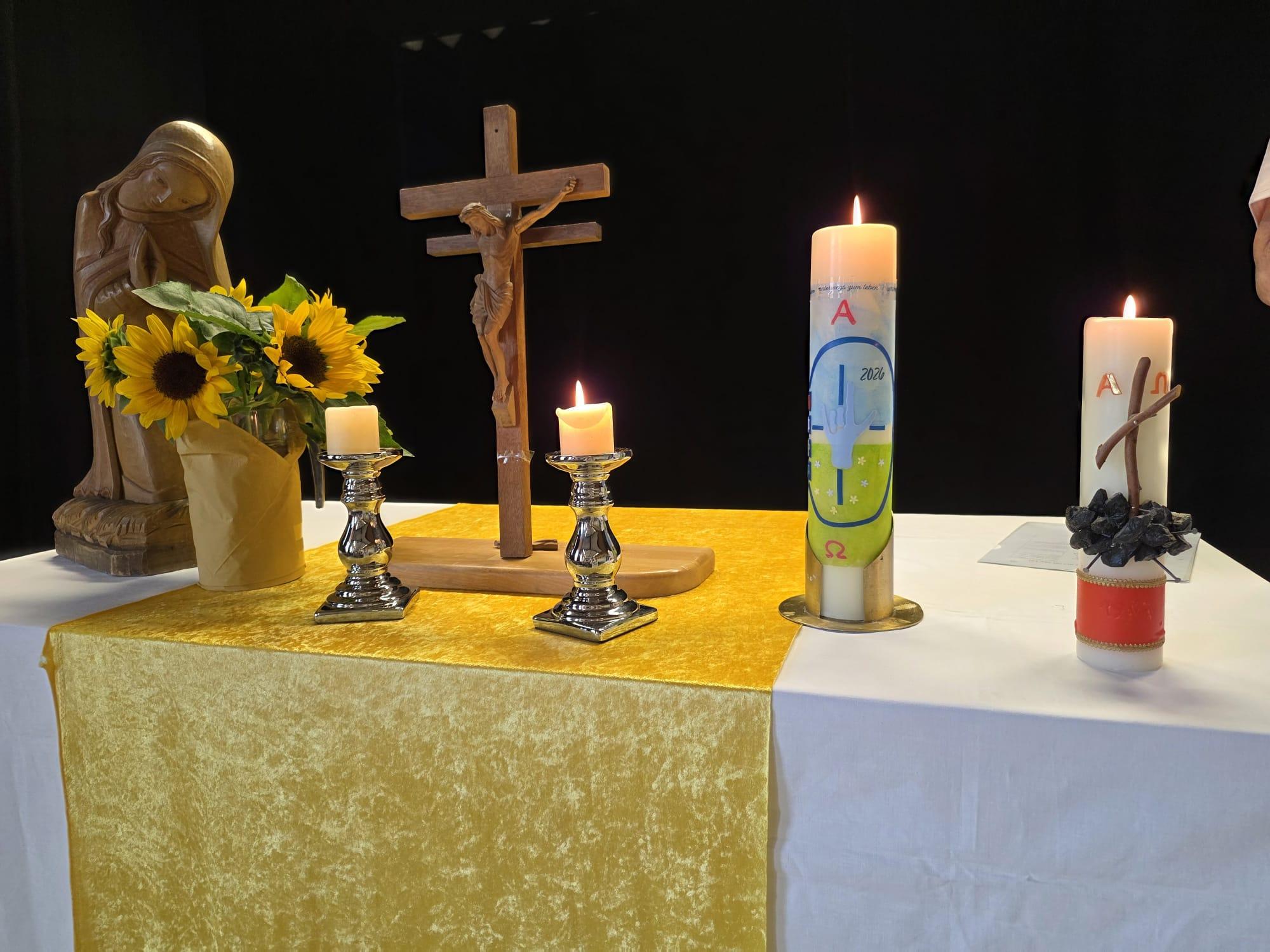 Kirchlich geschmückter Altar mit Kruzifix, Marienfigur, Sonnenblumen und brennenden Kerzen vor dunklem Hintergrund.
