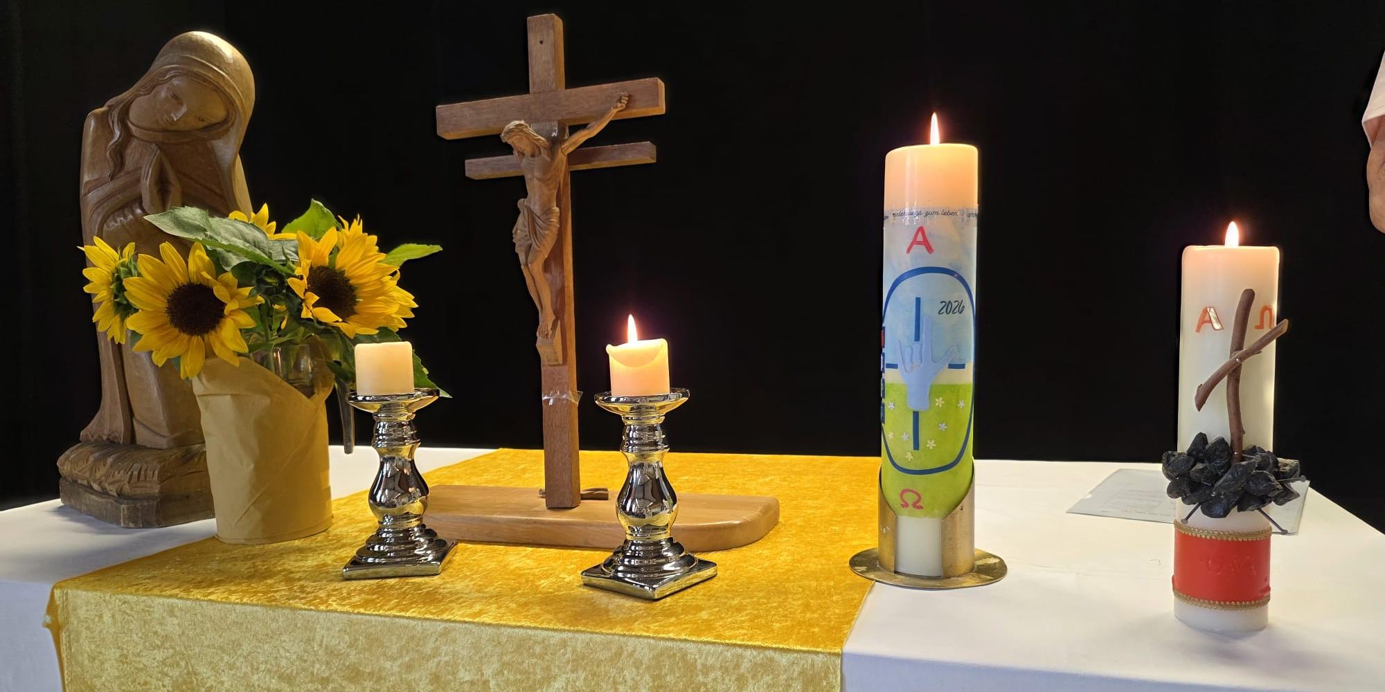 Kirchlich geschmückter Altar mit Kruzifix, Marienfigur, Sonnenblumen und brennenden Kerzen vor dunklem Hintergrund.