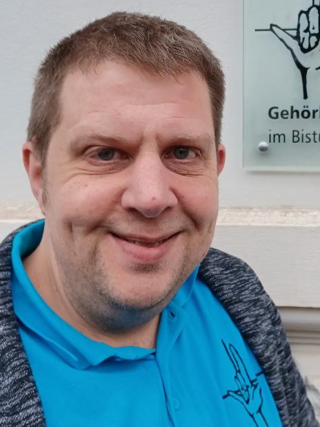Porträt einer Person vor einer Informationswand der Gehörlosenseelsorge im Bistum Trier.